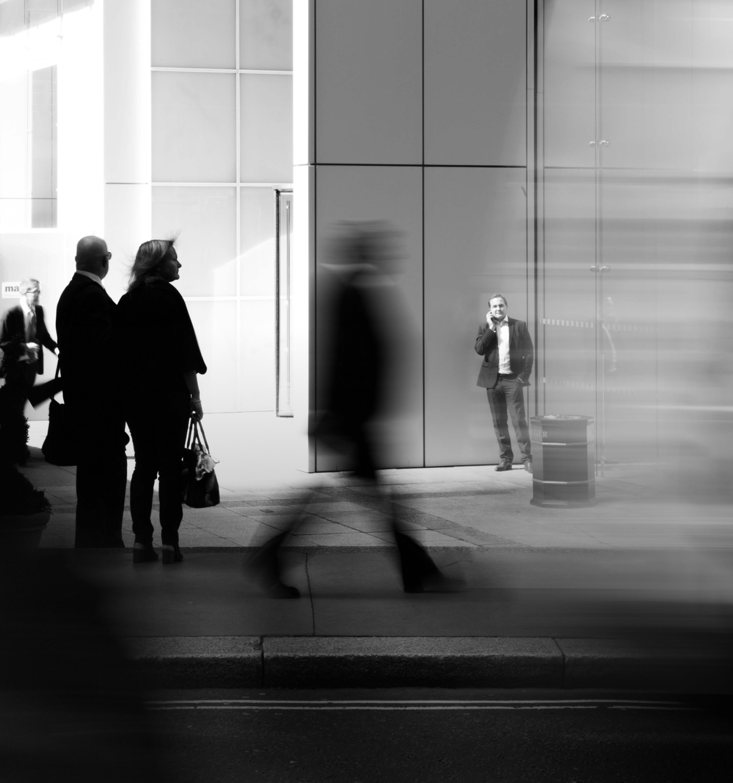 Black and white image of man standing in front of a building, talking on his phone.